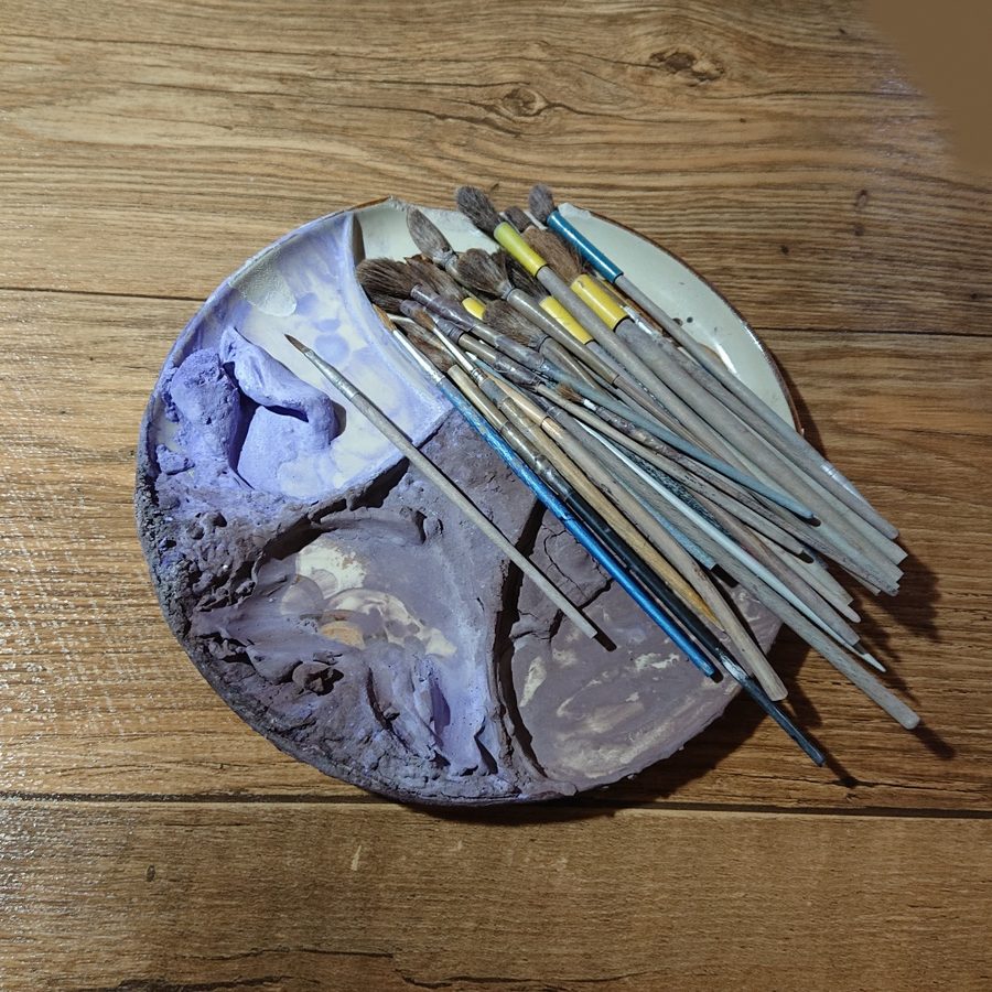 Runde Malerpalette mit angerührten Oxidfarben und einem Bündel Pinsel auf der Werkbank